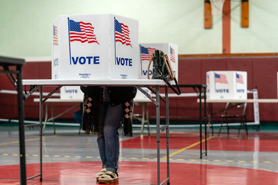 A person stands at a voting booth inside a gymnasium, with a purse on the table and "VOTE" signs displaying American flags on the booths.