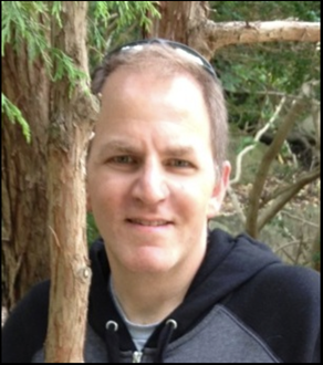 A man with short hair wearing a black hoodie stands outdoors near a tree, with greenery in the background.