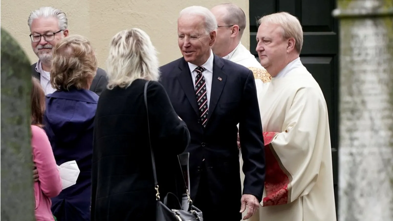 A group of people stand outside, including a man in a suit with a US flag pin, conversing with others, while a man in religious vestments stands nearby.