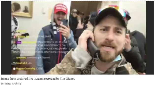 A man in a camouflage jacket speaks into a phone while others, some wearing hats and flags, gather closely behind him indoors.