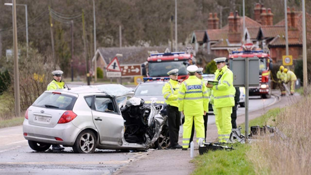 A damaged silver car is on the roadside with police officers and emergency crews at the scene of a traffic accident. Fire trucks are visible in the background.
