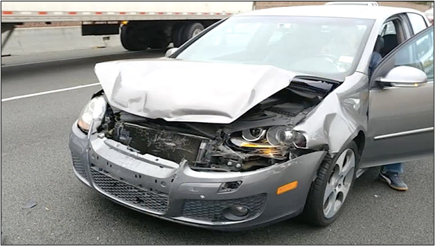 A silver car with severe front-end damage is stopped on the road, with the driver's door open and a truck visible in the background.