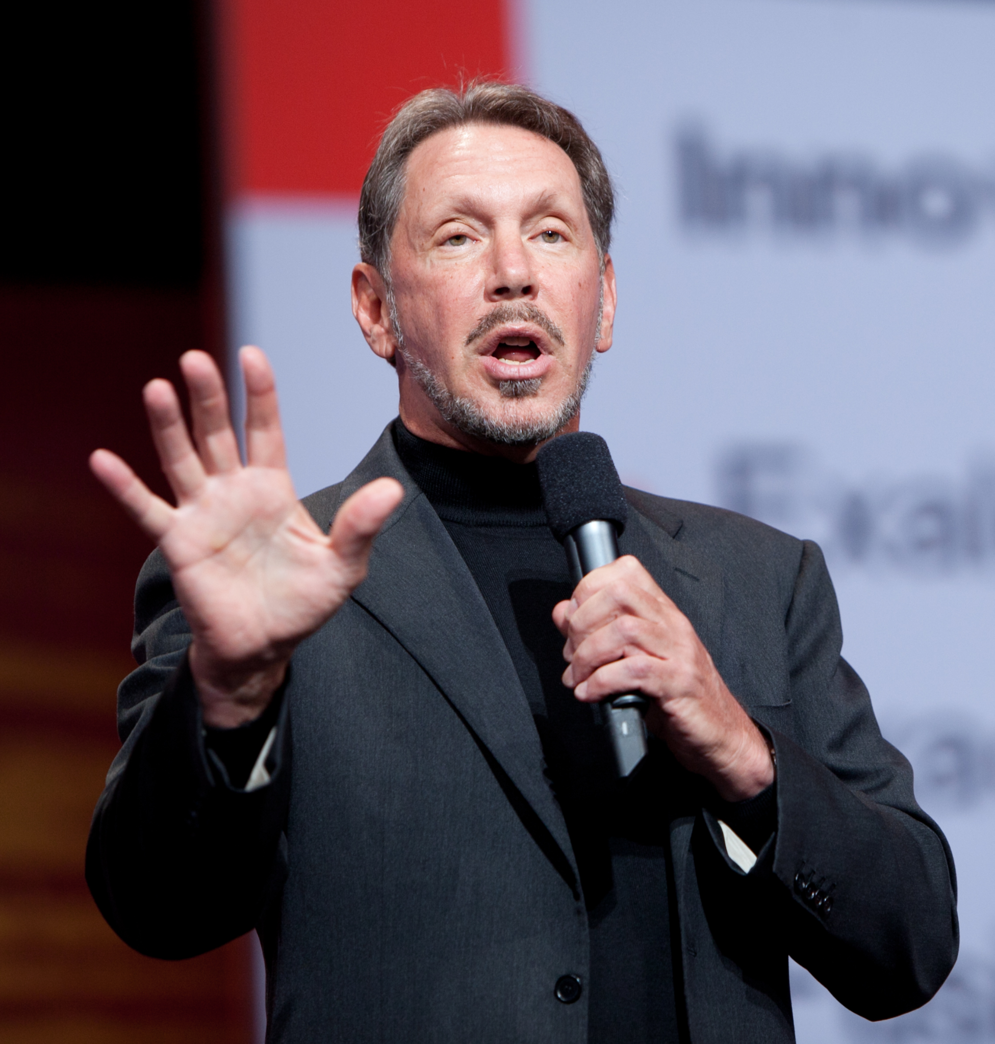 A man in a suit holding a microphone speaks on stage, gesturing with his left hand in front of a blurred presentation screen.