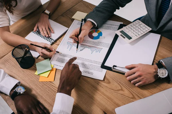 Three individuals are collaborating over a desk filled with documents, a graph, a notepad, sticky notes, and a calculator. They are discussing and pointing at the documents.
