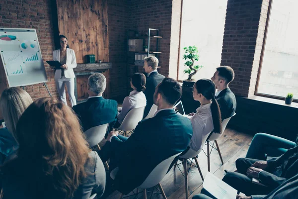 A person in a white suit presents graphs to an audience in a brick-walled room. The audience sits and listens attentively, some taking notes on laptops.