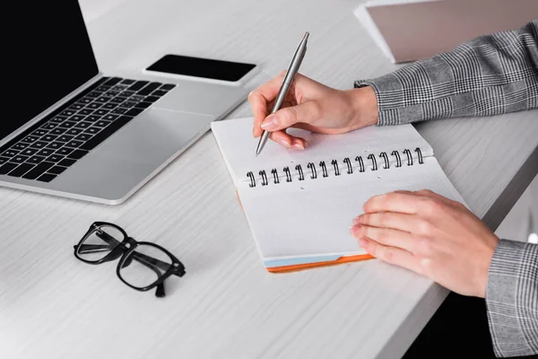Person in a plaid jacket writing in a spiral notebook on a white desk next to a laptop, smartphone, and a pair of glasses.