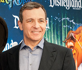 A man in a suit standing in front of a Disneyland Resort promotional backdrop featuring Beauty and the Beast artwork.