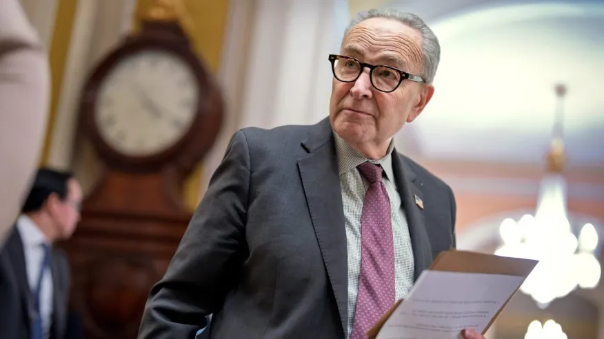 An older man in a suit and tie holds papers, standing indoors near a large clock and chandelier.