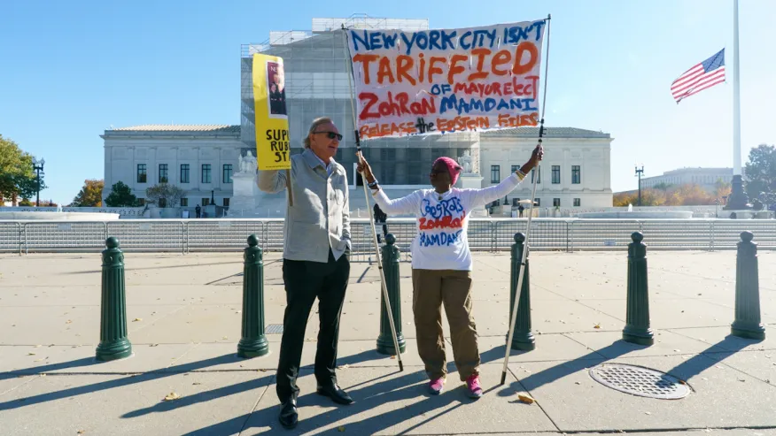Two people stand outside a government building holding protest signs; one sign references New York City taxes and mandates, and the other supports Rudy Giuliani.