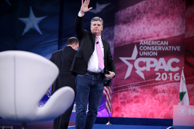 A man in a suit and pink tie waves while standing on stage at the 2016 American Conservative Union CPAC event, with a large backdrop and an empty chair visible.