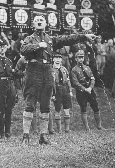 A man in a uniform gives a Nazi salute at an outdoor rally, with others in uniform and banners with swastikas visible in the background.