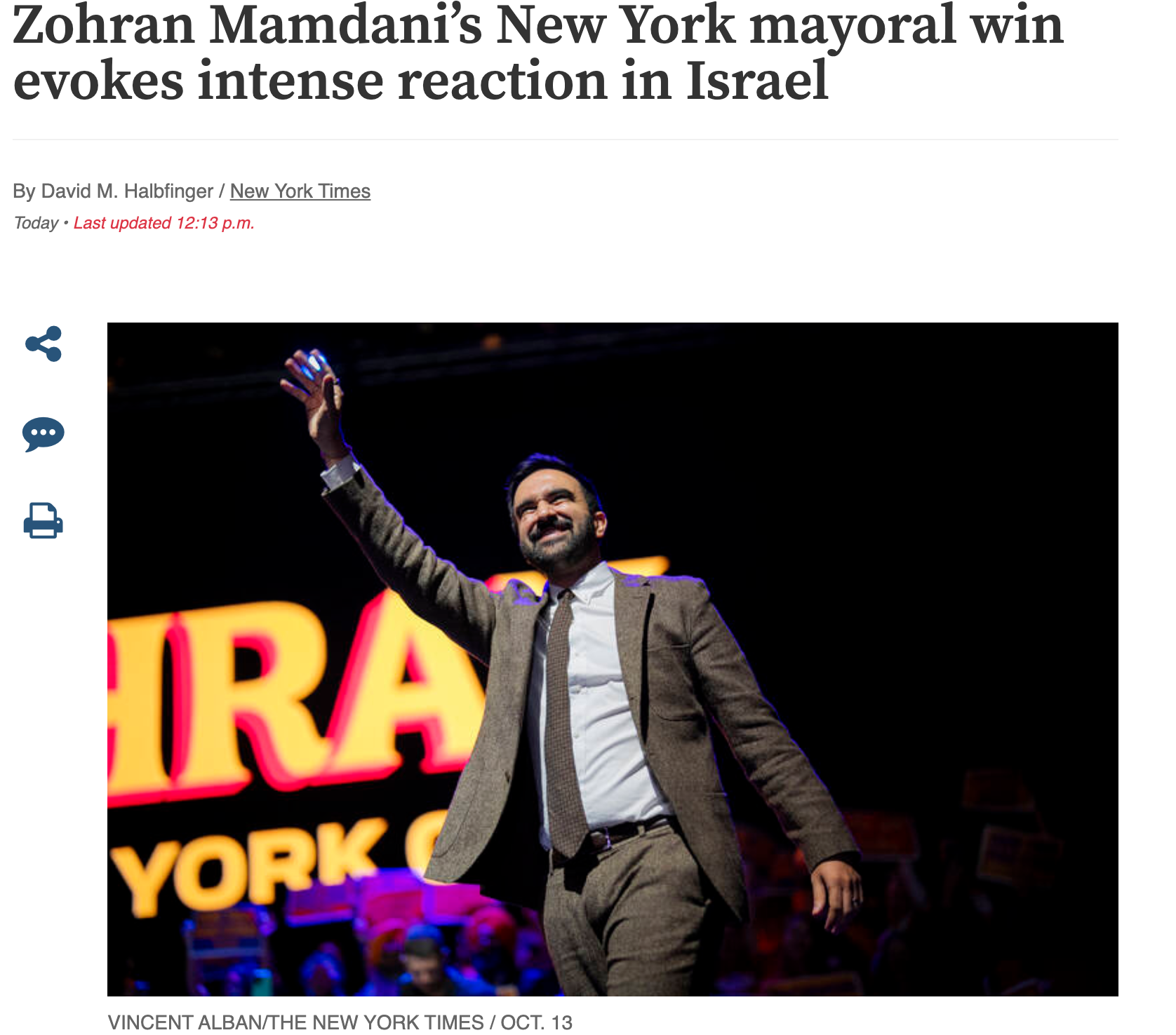A man in a suit stands on stage, smiling and waving to an audience, with bright lights and large text in the background.