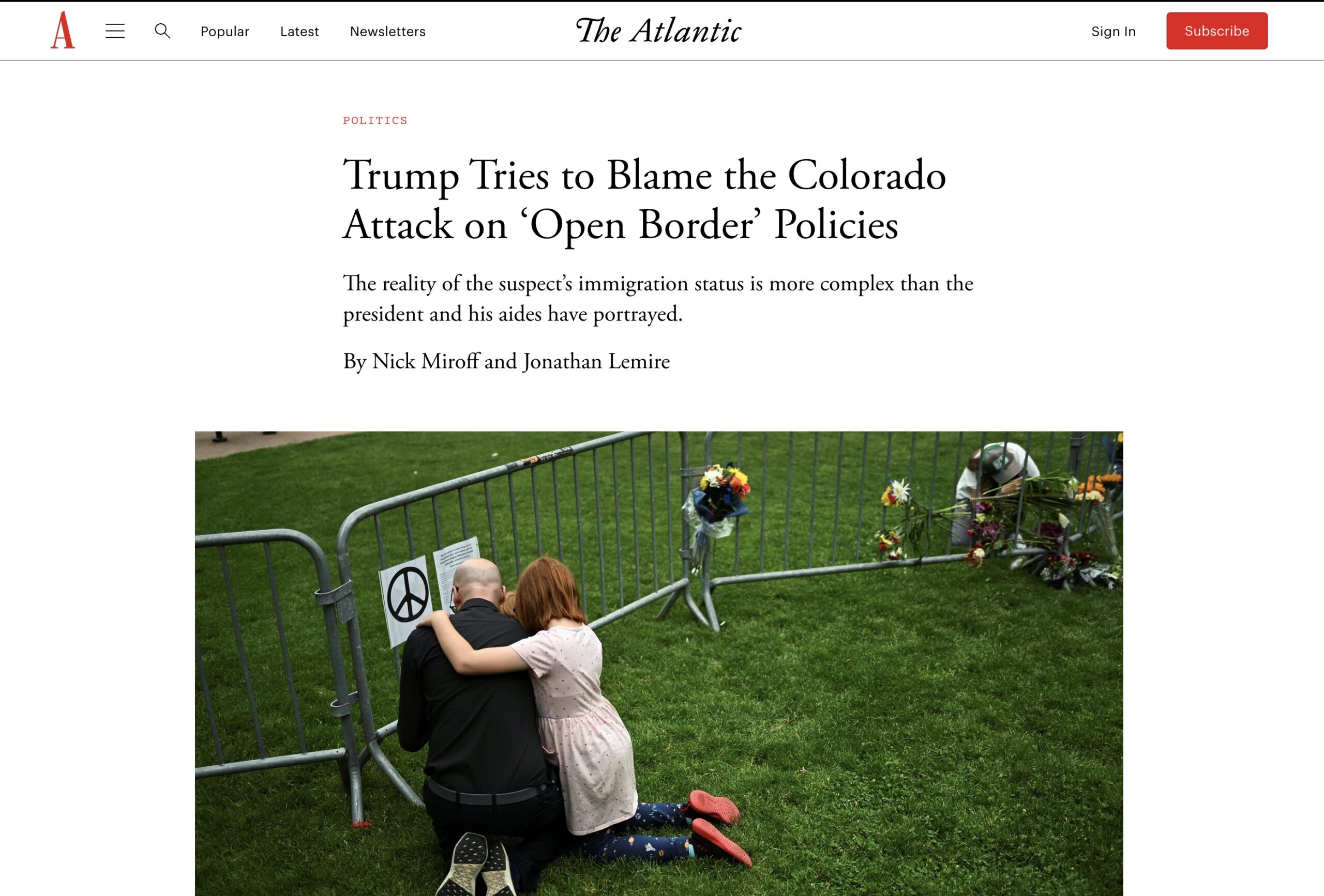 Two people kneel and embrace at a memorial with flowers and photos by a fence on a grassy area, mourning victims of an attack.