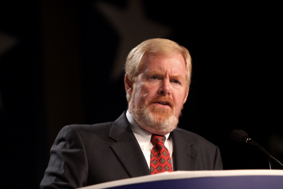 A middle-aged man with a light beard and suit speaks at a podium with a microphone, set against a dark background featuring blurred star shapes.
