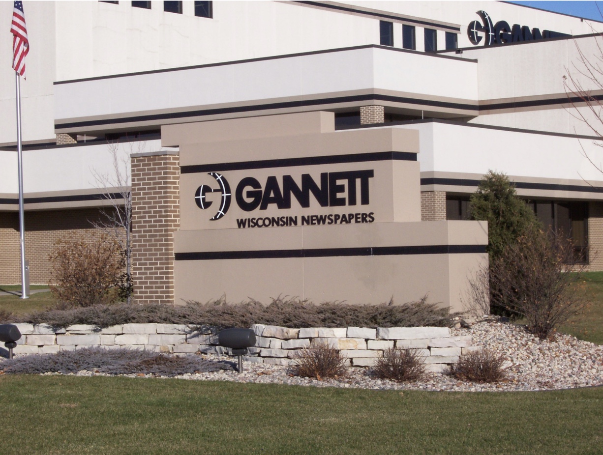 A sign outside an office building reads "Gannett Wisconsin Newspapers" with the Gannett logo, surrounded by landscaping and grass.