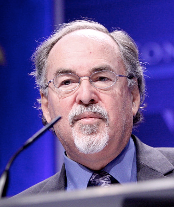 An older man with gray hair, glasses, and a goatee is wearing a suit and tie, speaking at a podium against a blurred blue background.