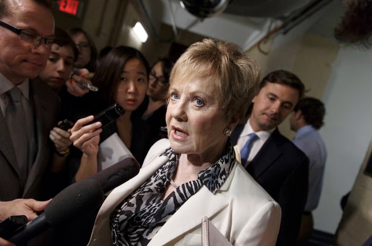 A woman in a white blazer speaks to reporters holding microphones and recorders in a crowded hallway, with people standing and listening around her.