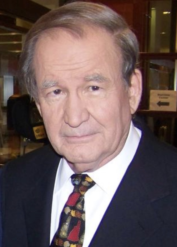 A man with short gray hair in a dark suit, patterned tie, and white shirt stands indoors, looking at the camera with a neutral expression.