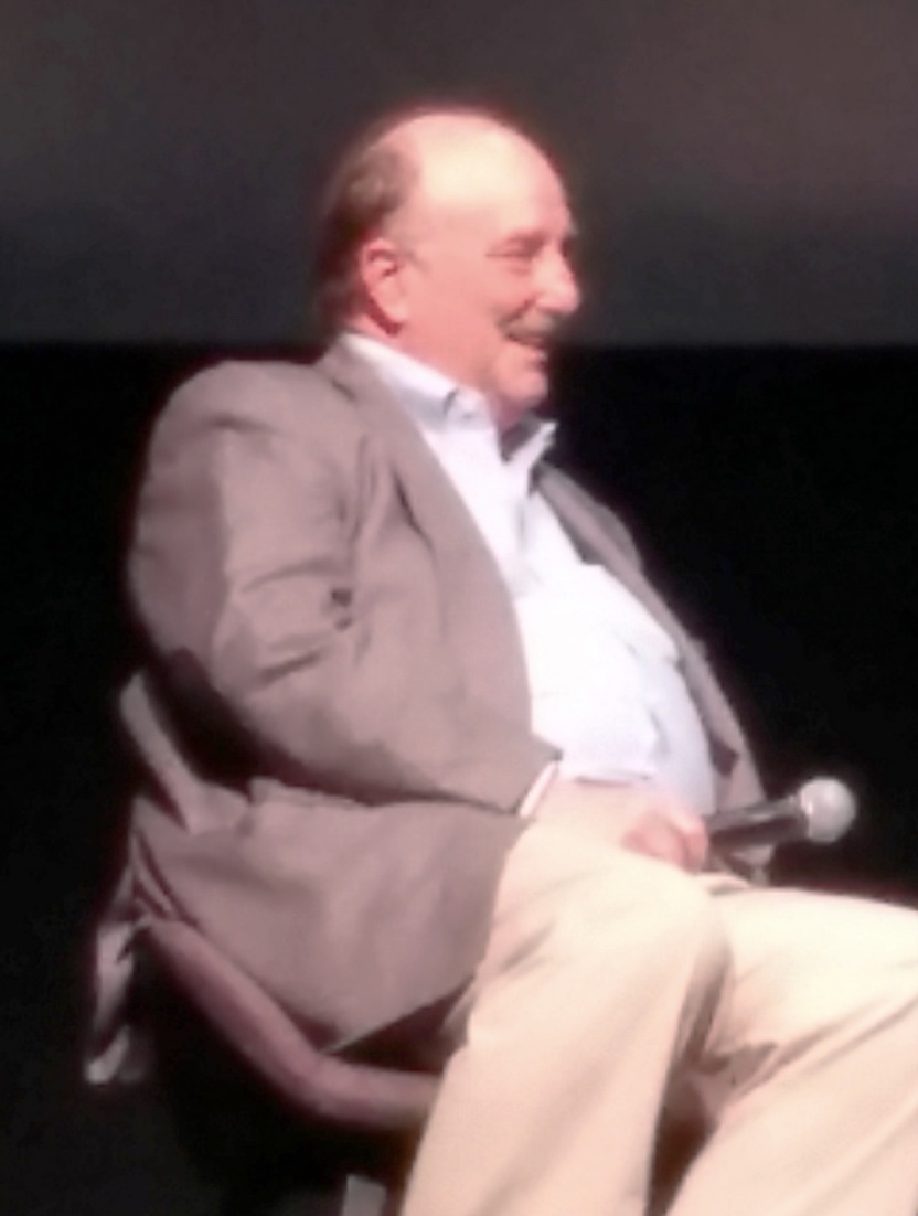 A man with a mustache sits on a chair, holding a microphone, wearing a gray blazer, light blue shirt, and beige pants against a dark background.
