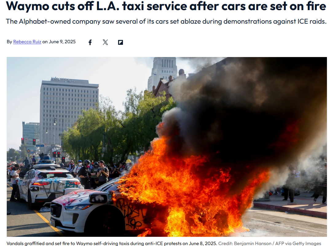 A Waymo self-driving car is engulfed in flames on a city street during a protest, with emergency responders and bystanders nearby.