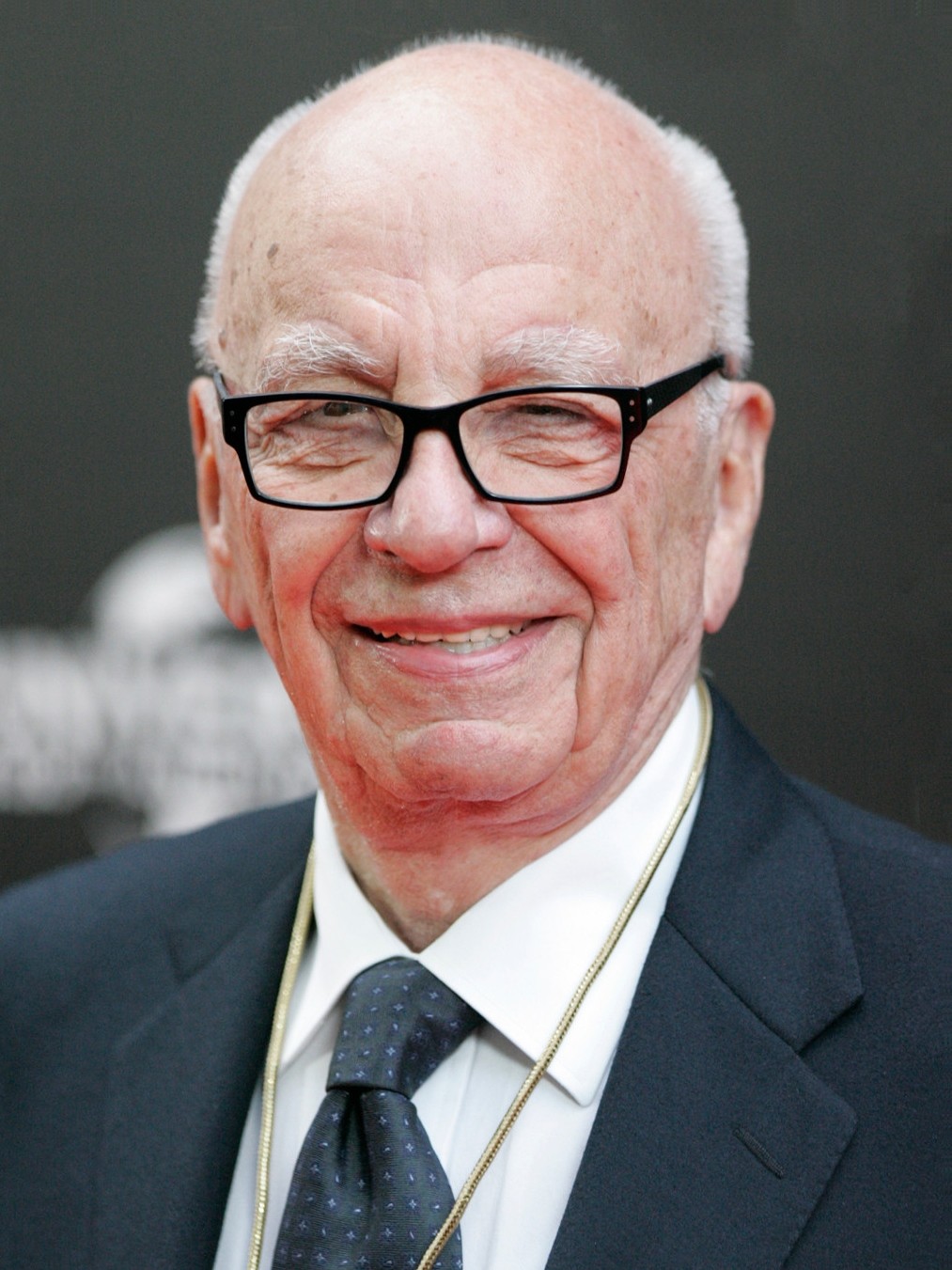 An older man with glasses, wearing a dark suit, white shirt, and polka dot tie, smiles at the camera against a dark background.