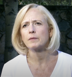 A woman with short blonde hair and a white shirt looks ahead with a serious expression; stone wall and trees are visible in the background.