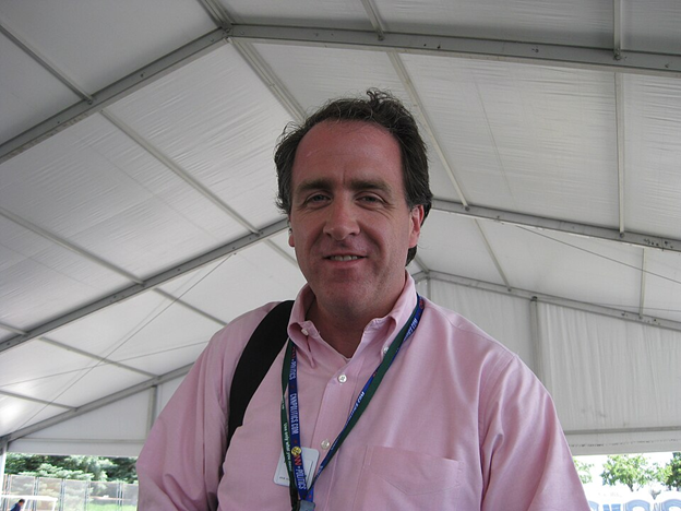 A man wearing a light pink shirt and a lanyard stands under a large white event tent, carrying a black shoulder bag.
