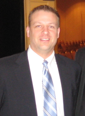 A man in a suit and striped tie stands indoors, facing the camera and smiling.