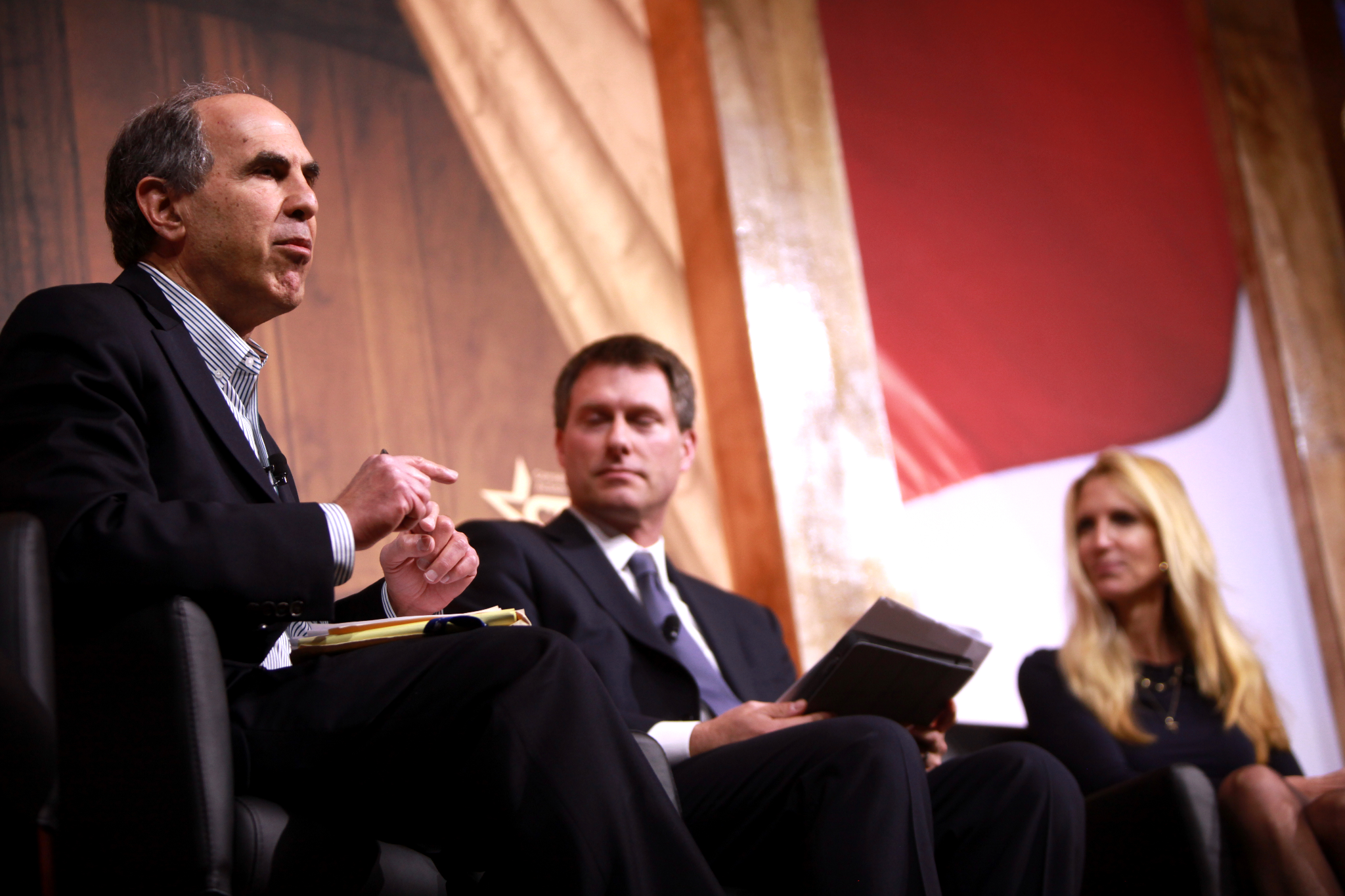 Three people in business attire sit on a stage, engaged in a panel discussion with two men holding notes and a woman listening attentively.