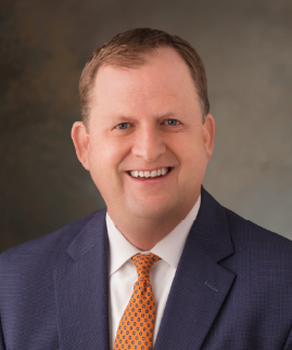 A man in a blue suit, white shirt, and orange patterned tie smiles at the camera against a neutral background.