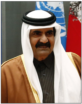A man in traditional Middle Eastern attire stands in front of flags, including the United Nations flag, and looks slightly to the side.