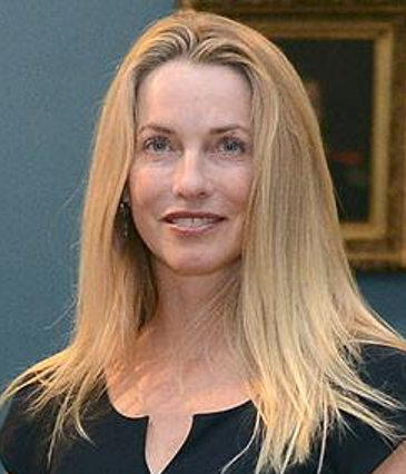 A woman with long straight blonde hair, wearing a black top, stands indoors in front of a framed painting.
