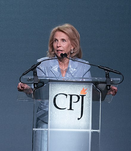 A woman in a blue blazer speaks at a podium with the initials "CPJ" and a flame logo.