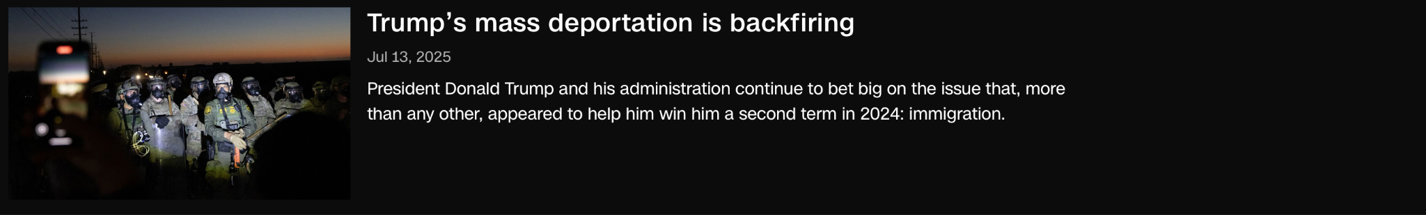 A group of uniformed officers stands at night near a fence, illuminated by bright lights; a border wall and vehicles are visible in the background.