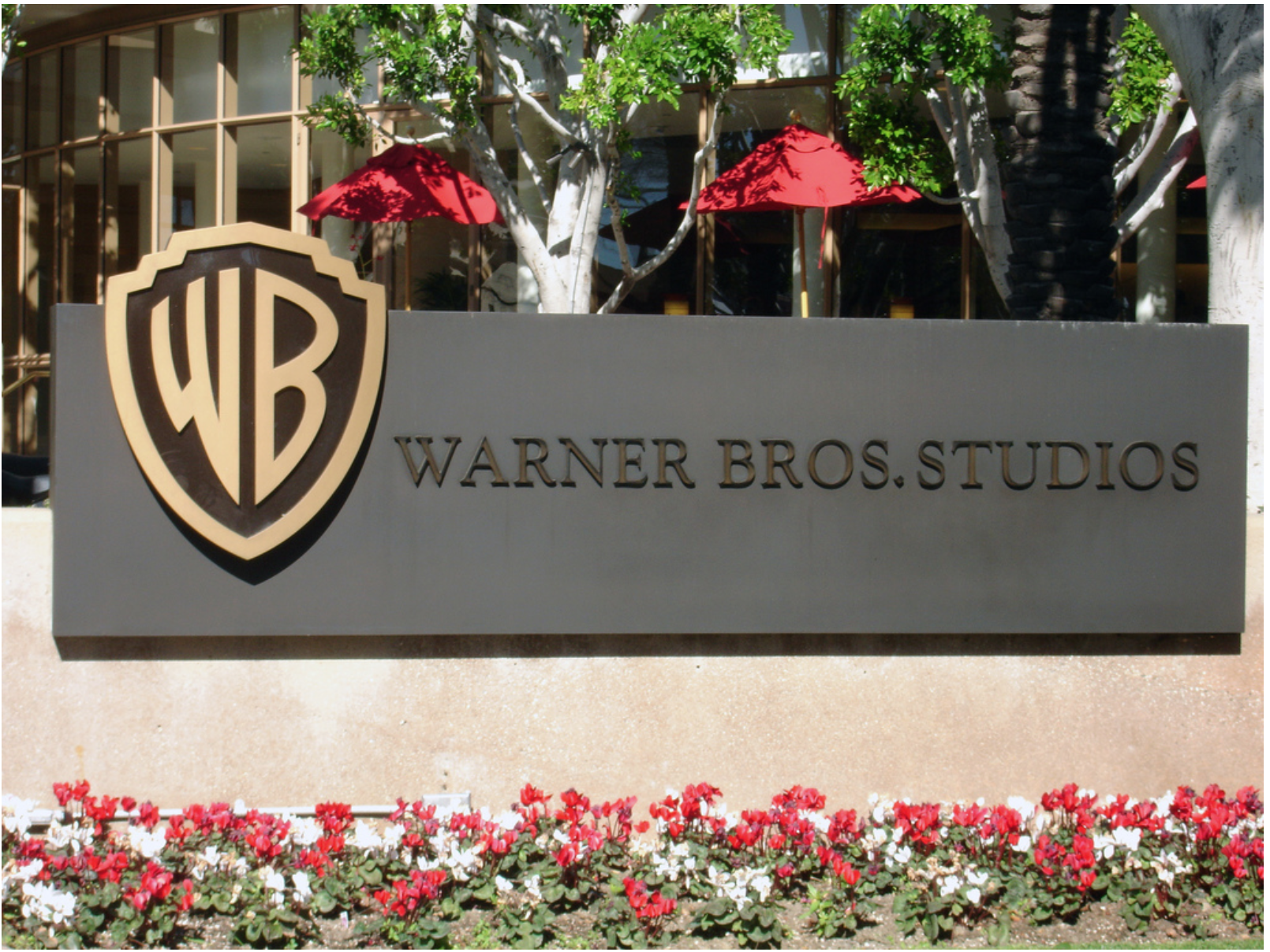 A sign with the Warner Bros. shield logo and the text “Warner Bros. Studios,” with red and white flowers and red umbrellas in the background.