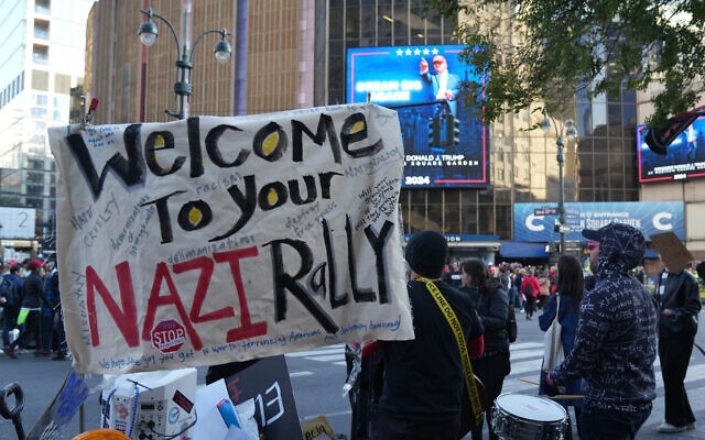 Protesters with a sign reading "Welcome to your Nazi rally" at an outdoor demonstration, with people and a large building in the background.