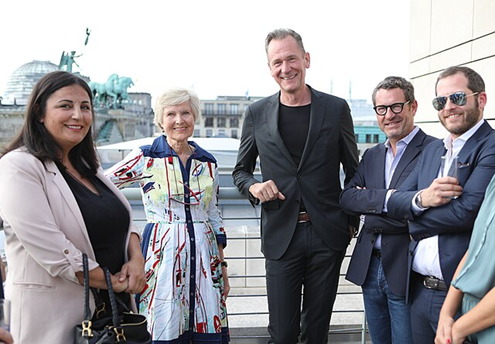 Five adults, three men and two women, standing and smiling together on an outdoor terrace with city buildings in the background.