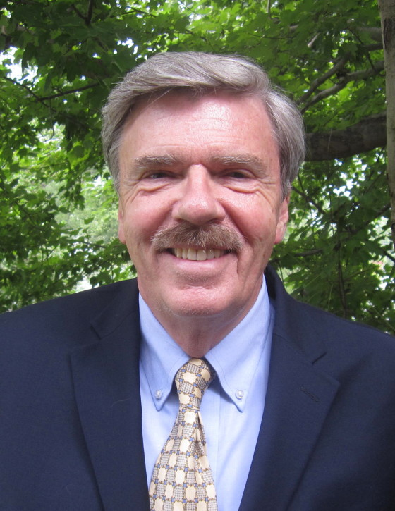 A man in a suit and patterned tie smiles at the camera while standing outdoors in front of leafy green trees.