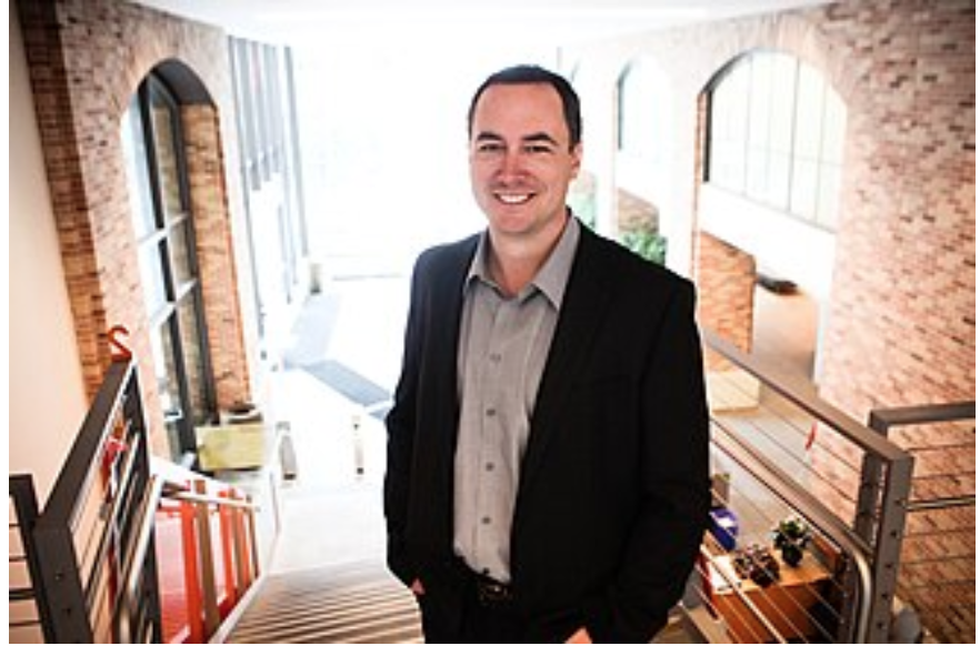A man in a dark suit stands on a staircase inside a building with brick walls and large windows, smiling at the camera.