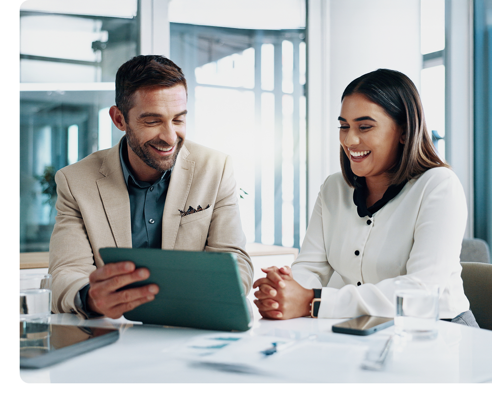 Two business professionals sit at a table, smiling and looking at a tablet together in a modern office setting.