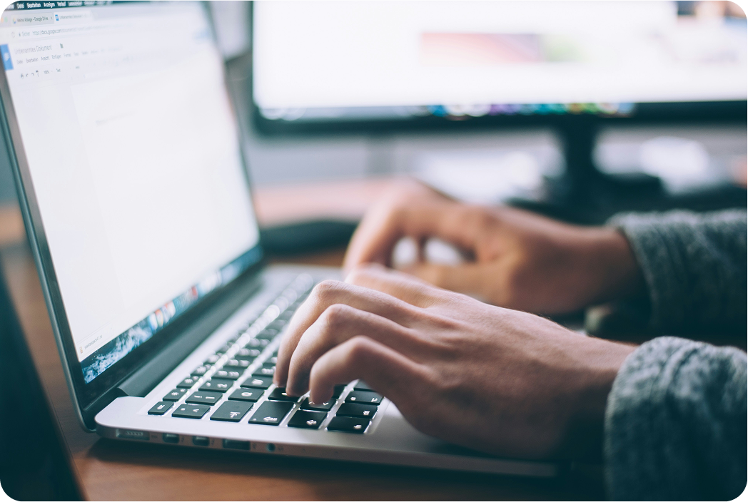 Close-up of a person typing on a laptop keyboard, with a blurred computer monitor and workspace visible in the background.