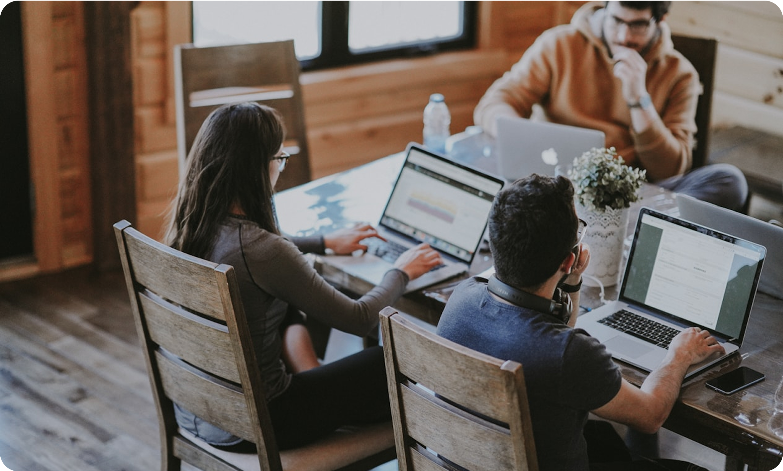 Three people work on laptops at a wooden table in a cozy room. A plant and a water bottle are on the table.