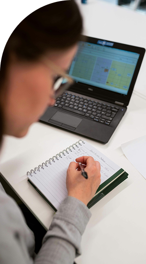 A person writes in a spiral notebook at a desk, with an open laptop displaying a spreadsheet in the background.