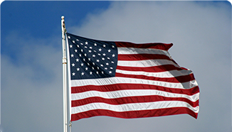 The United States flag with 50 stars and 13 stripes is flying on a flagpole against a partly cloudy sky.