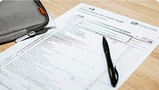 A black pen rests on a completed IRS Form 1120 tax document, with a gray zippered case nearby on a wooden surface.
