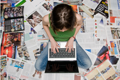 A person sits cross-legged on the floor, typing on a laptop, surrounded by scattered newspapers and magazines.