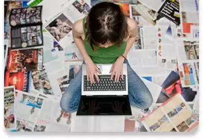 A person sits cross-legged on the floor, typing on a laptop, surrounded by scattered open magazines and newspapers.