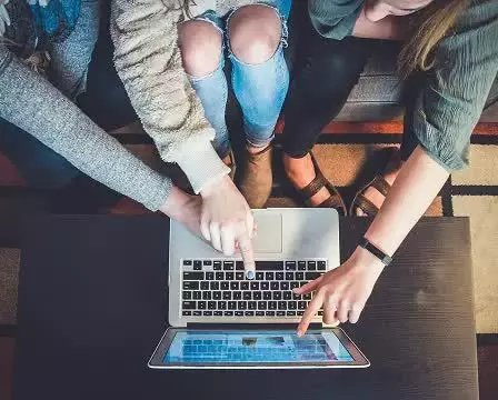 Three people sitting together point at the screen of an open laptop on a table, viewed from above.