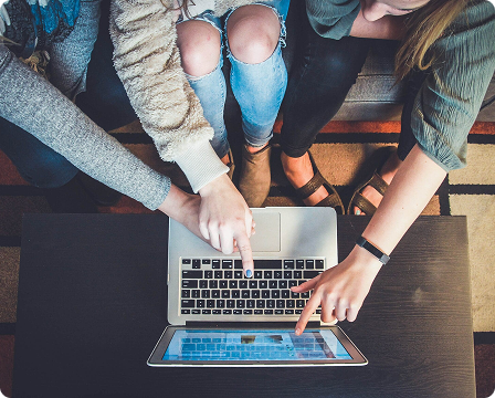 Three people sitting on a couch point at the screen of a laptop on a coffee table, viewing something together.
