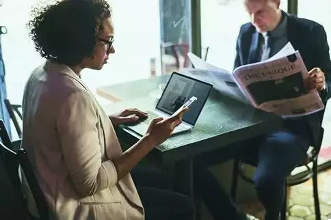 A person sits at a table using a smartphone and laptop while another person nearby reads a newspaper in a cafe setting.